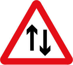 UK two-way traffic straight ahead warning sign: a red-bordered triangle with two black arrows, one pointing up and one pointing down